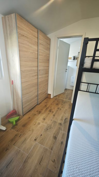 Ein Zimmer mit einem Schrank, einer offenen Tür und einem Hochbett. Bodendielen sind sichtbar.