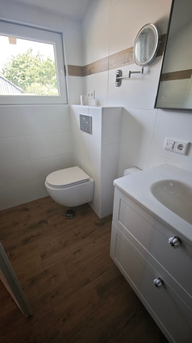 Helles Badezimmer mit Toilette, Waschbecken und Fenster. Boden aus Holzoptik.