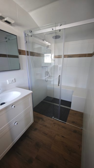 Moderne Dusche mit Glastür in einem hellen Badezimmer. Weißer Waschtisch und Holzboden.